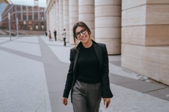 American brunette young businesswoman in black suit looks at camera happily smiles walking in street