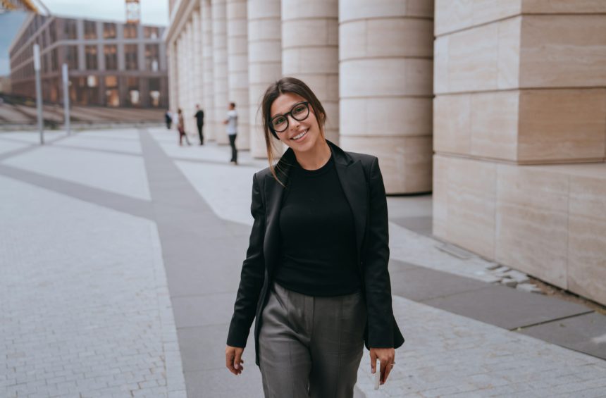 American brunette young businesswoman in black suit looks at camera happily smiles walking in street