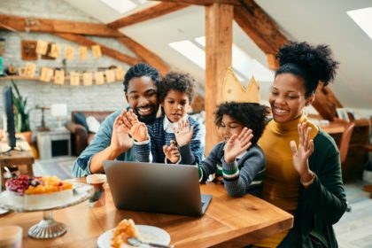 Familie afro-americană fericită care sărbătorește ziua de naștere a fiicei și făcând cu mâna cuiva în timpul unui apel video pe laptop de acasă.