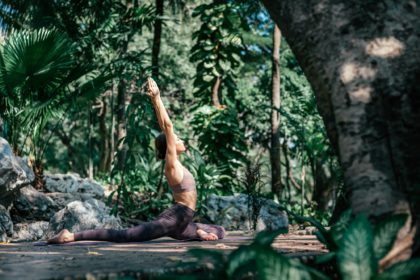 Fotografie pe toată lungimea unei femei caucaziene stând în poziție Pigeon Upright, Kapotasana în timp ce practică yoga în aer liber, într-o grădină.